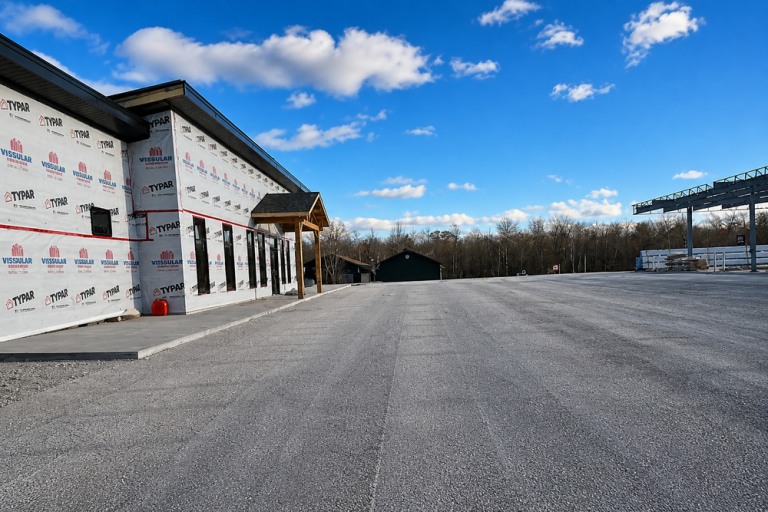 Wide view of a commercial asphalt paving project in summer, showing a newly paved parking area beside a building under construction with clear skies.