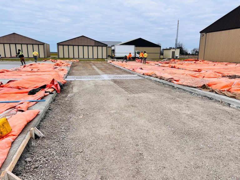 Commercial asphalt and concrete preparation site showing workers paving and preparing the foundation near industrial buildings.