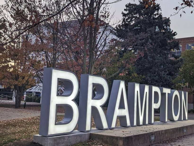 Large illuminated “BRAMPTON” sign in a public square surrounded by trees, walkways, and nearby buildings at dusk.